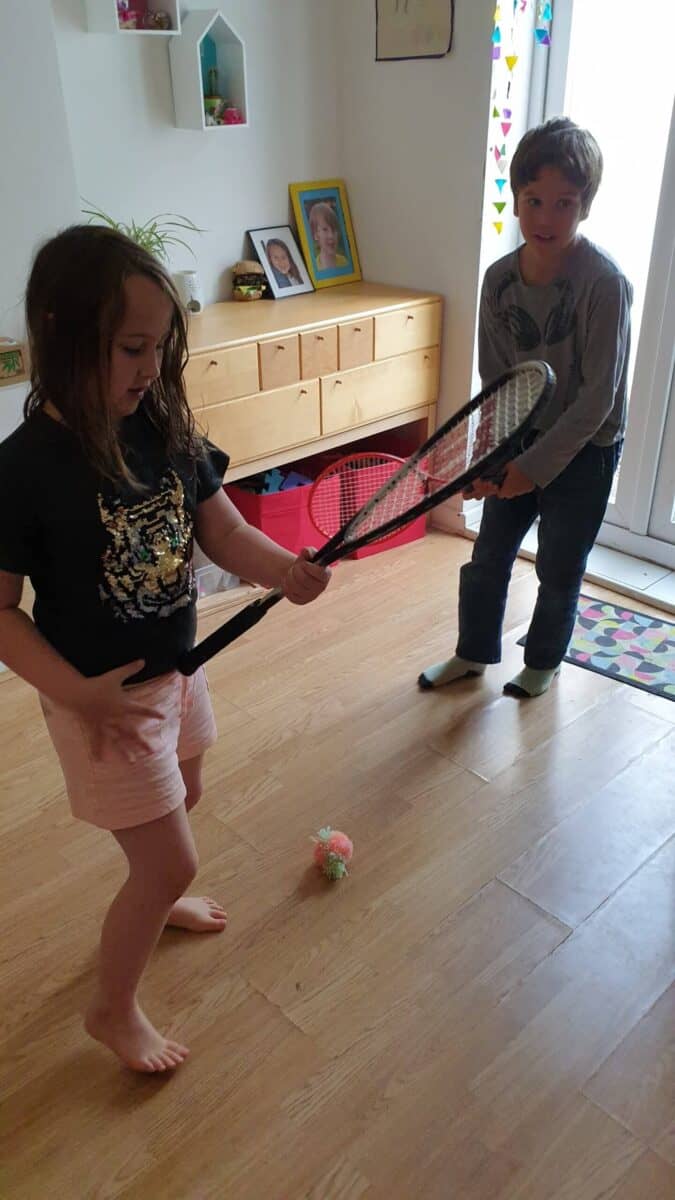 An image of two children, a ball on the floor, both holding tennis rackets.