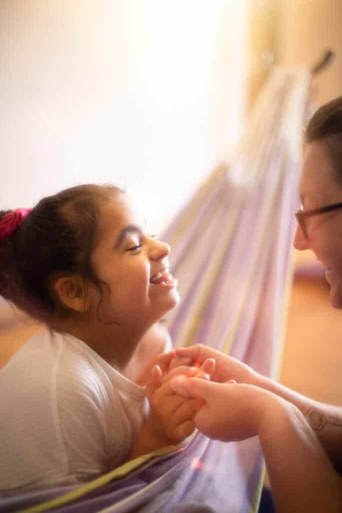The images is of a young girl holding hands with an adult, sat in a hammock smiling at each other.