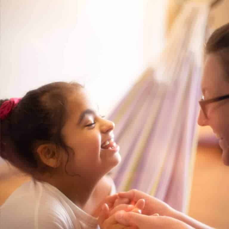 Image is of a young girl smiling at a lady holding her hand whilst sat in a hammock.