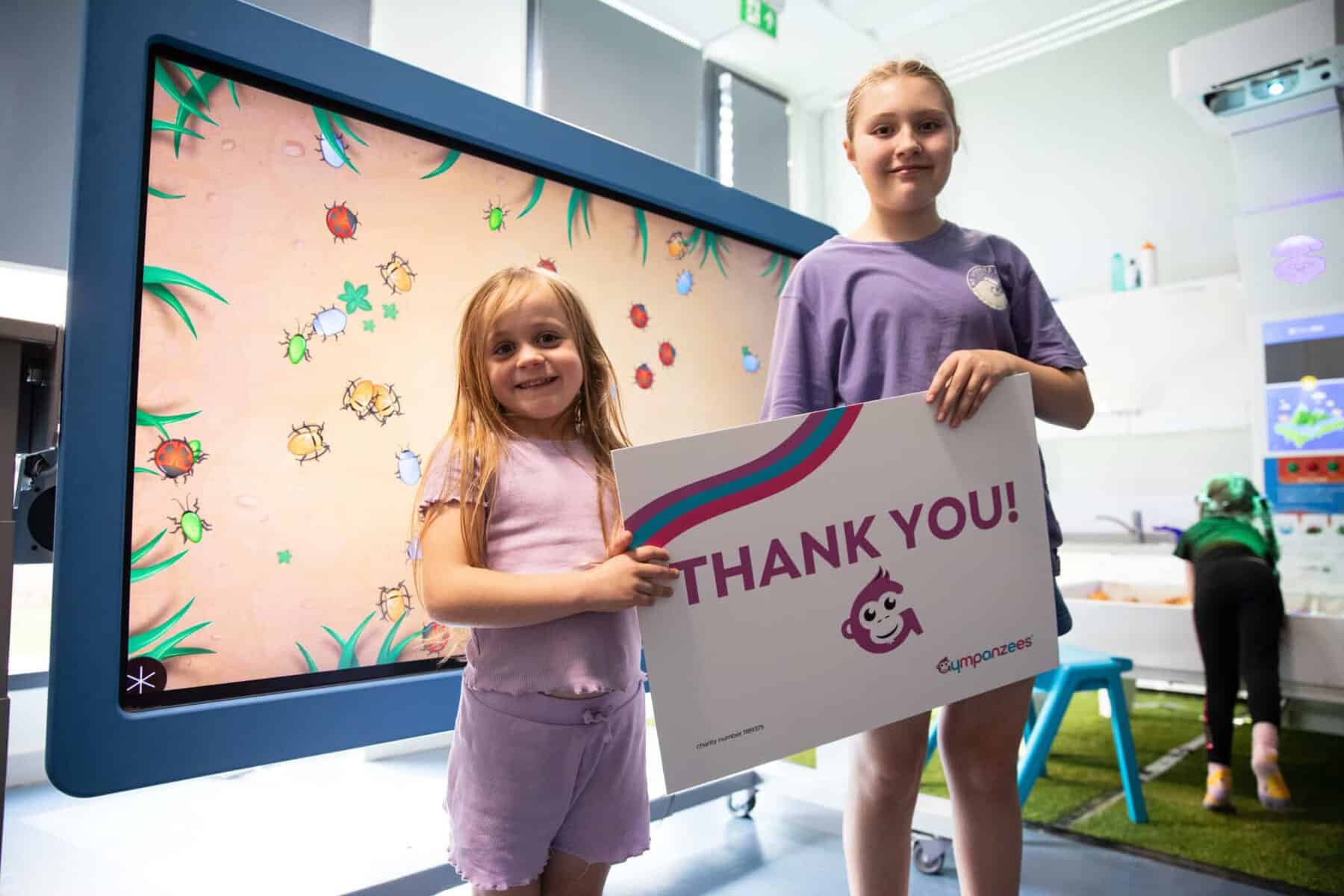 Two girls at Gympanzees holding a thank you sign, smiling into the camera, with an interactive large screen in the background.