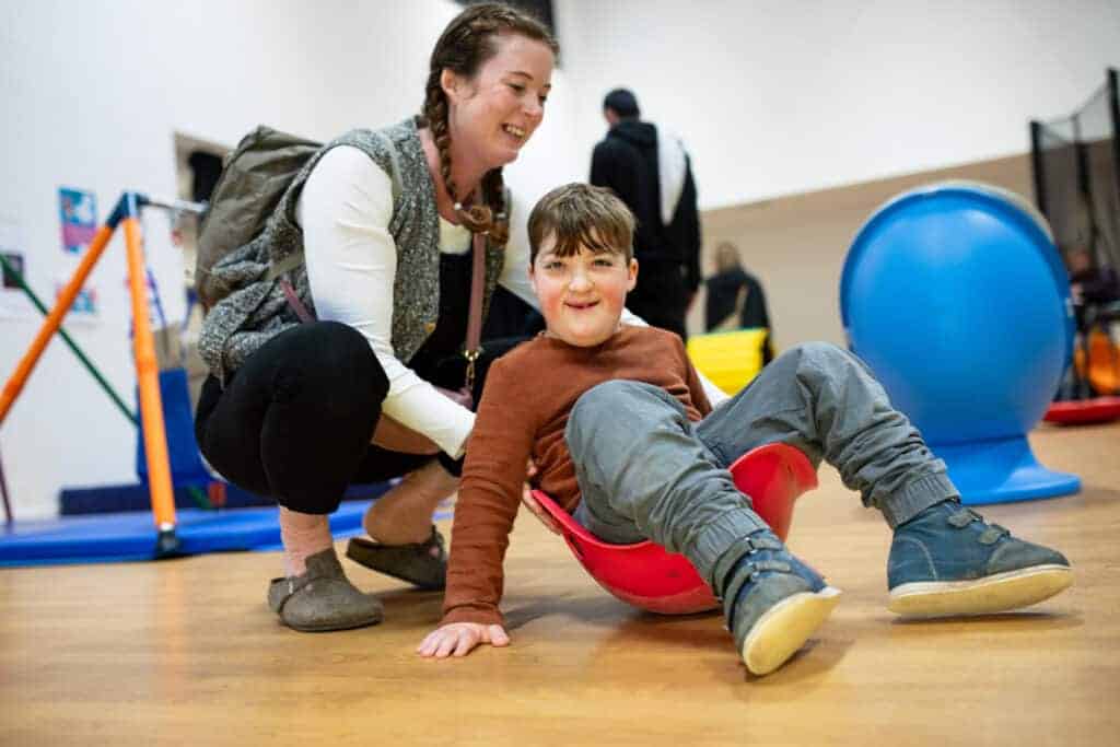 The circular image is of a young child sat in a spinning top toy, with an adult assisting behind them, with a gym hall background.