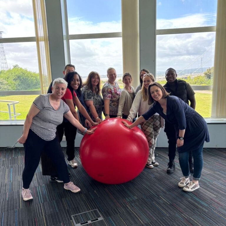 A group of people standing together outdoors around a large gym ball, each with one hand resting on it.