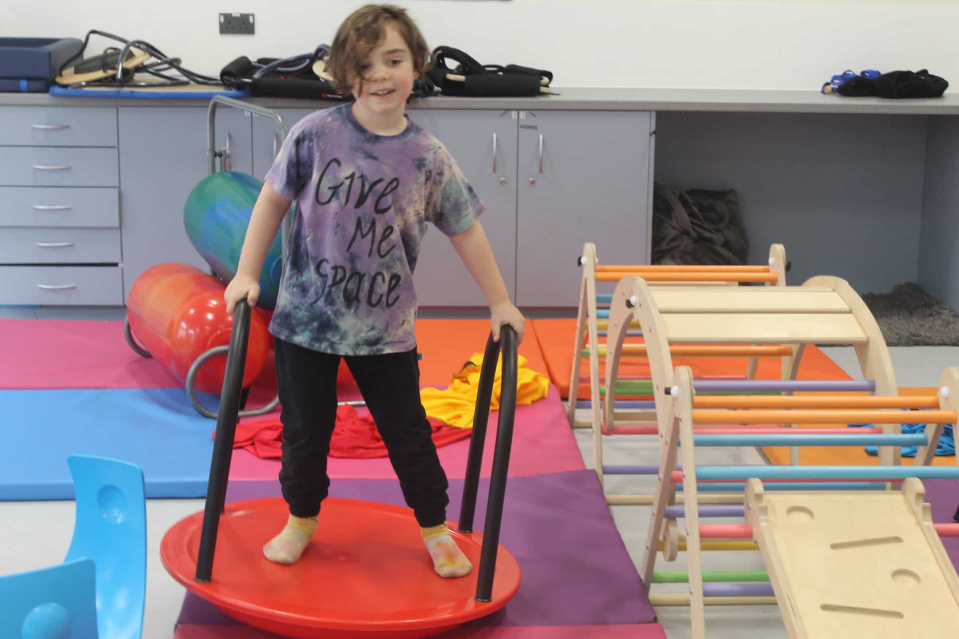 A child with a big smile stands confidently on a red balance board with handrails, wearing a colorful tie-dye shirt that reads 'Give Me Space.' The room is set up with vibrant mats, sensory equipment, and wooden climbing frames, creating an engaging and inclusive play environment