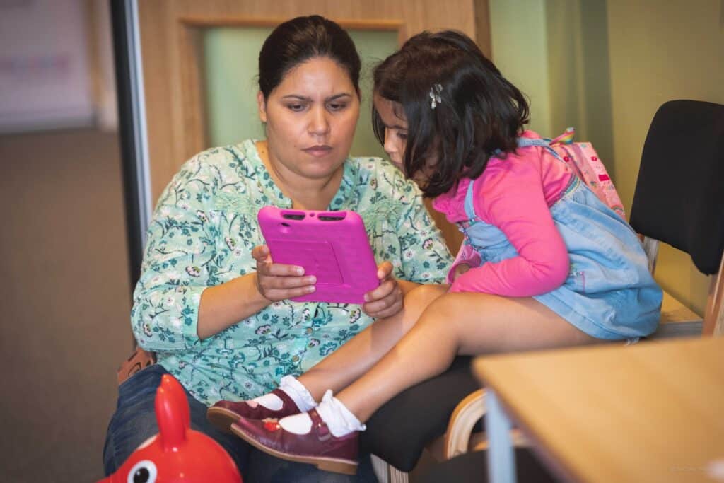 A woman and a young girl are sitting together, looking at a pink tablet. The woman, wearing a green floral top, holds the tablet while the girl, dressed in a pink long-sleeve shirt and denim dress with a backpack, leans in attentively. They are in an indoor setting with warm lighting.