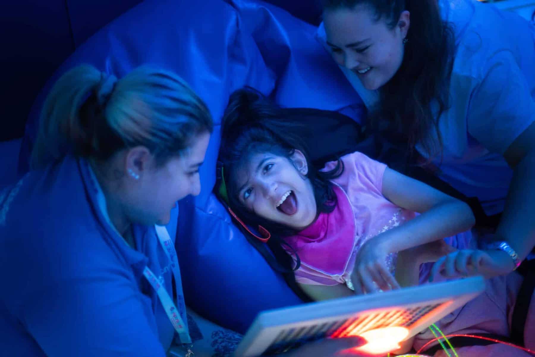 The image is of a two adults and a child in the middle all sat on a giant bean-bag in a darkly lit room, holding a marble board, with fibre optic lights behind.