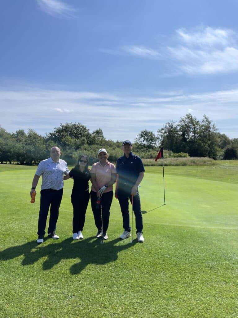 The image is of four people smiling, holding golf clubs on a green lawn, with a hole in the background due to play golf.