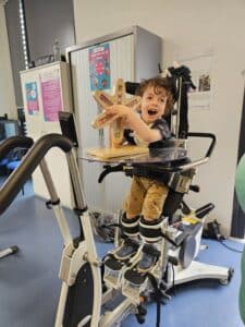 The image is of a young boy holding a sensory toy, in an assisted standing frame that simulates walking. This is with a classroom background.