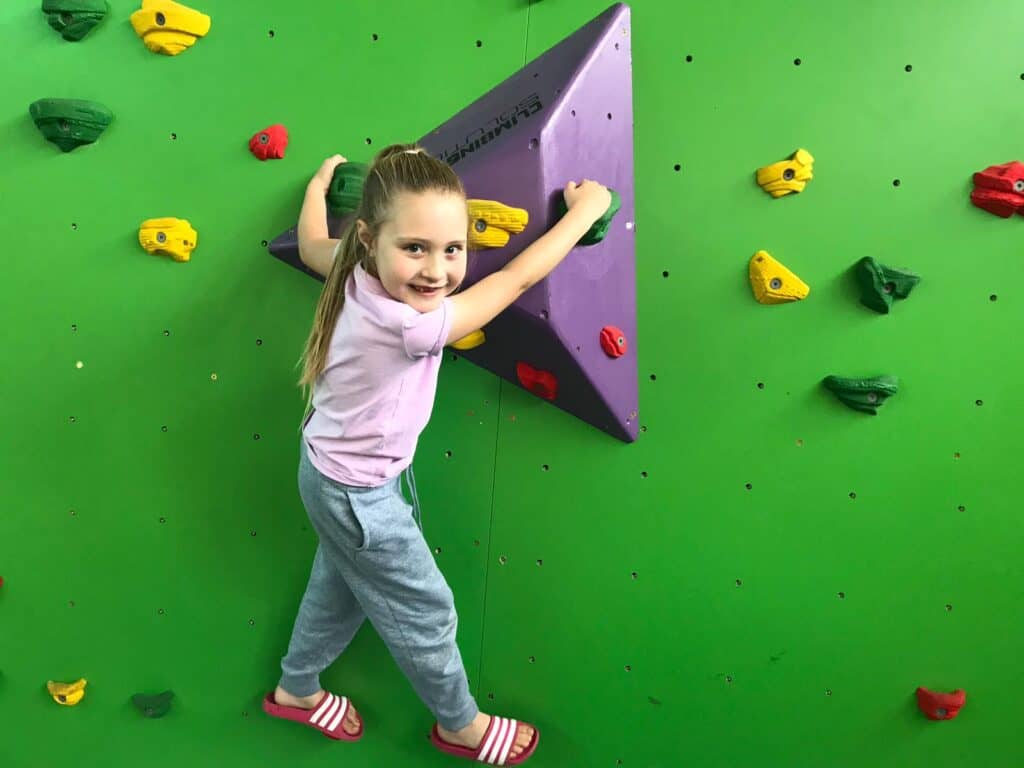 This circular image contains a child holding onto boulders on a boulder, climbing wall. They're turn to face the camera behind smiling.
