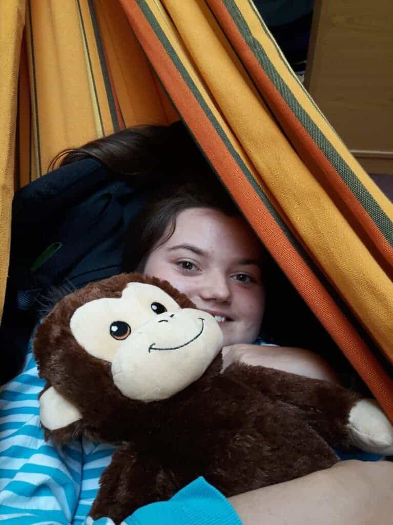The image is of a girl smiling from the shoulders up holding a cuddly toy monkey, in what appears to be a hammock that she is sitting in, in the background.