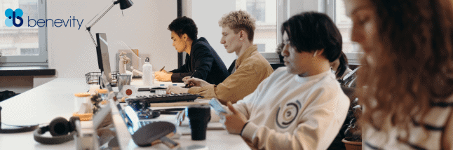 The image is from end of a long desk with multiple people sat at their desks looking at their computer screens in an office envrionment.