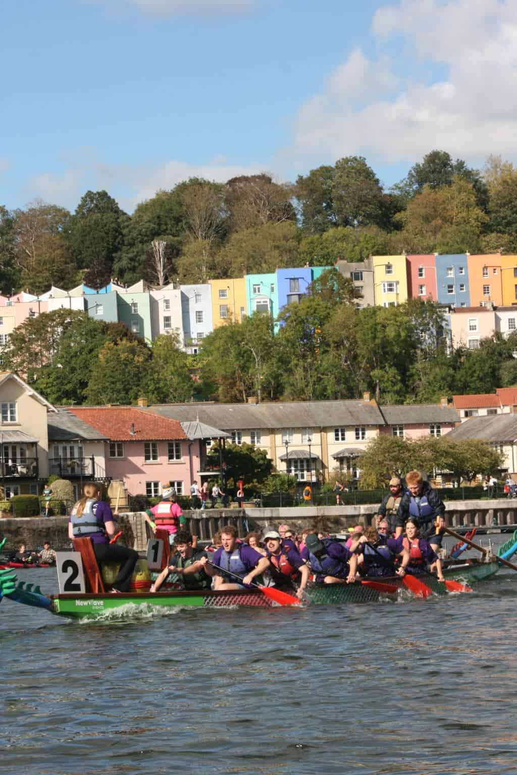 The image is of several people in a Dragon boat, all paddling, with one person sat at the end as a drummer taking part in a race on a river with a house and forestry background.