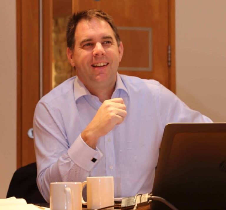 A person sat at his desk with laptop and mugs in front of him smiling