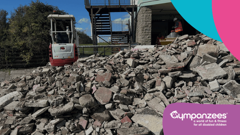 A pile of rubble in front of a building with a construction worker and a small excavator, under a clear sky. The logo in the corner indicates "Gympanzees, a world of fun & fitness for all disabled children.
