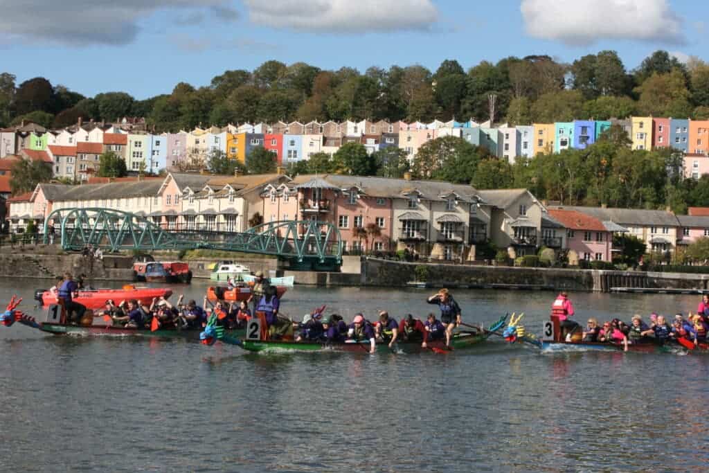 The image is of a large river with a cityscape in the background and several dragon boats on the water paddling taking part in a race.