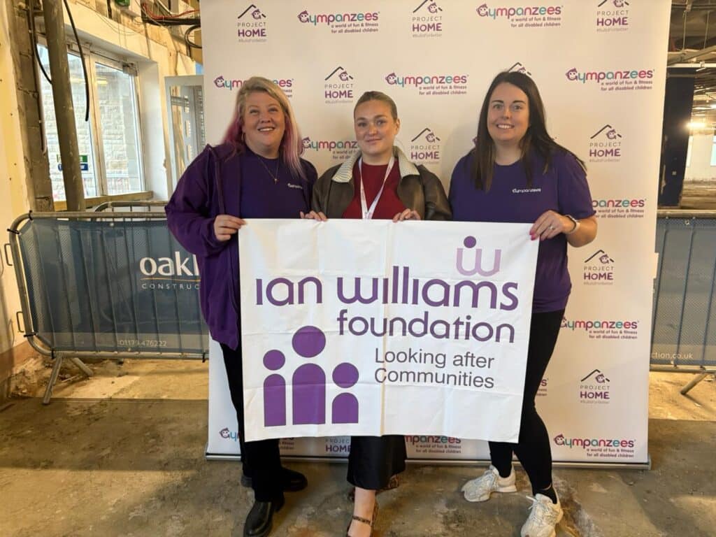 Three people stand smiling in front of an Ian Williams Foundation banner, holding a sign that says, “Looking after Communities.” They wear branded clothing, with the Gympanzees and Project Home Appeal logos visible in the background.