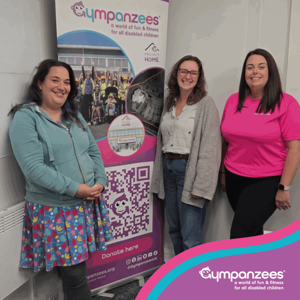 Three individuals standing beside a promotional banner for Gympanzees. The banner features a logo, QR code, and website URL.