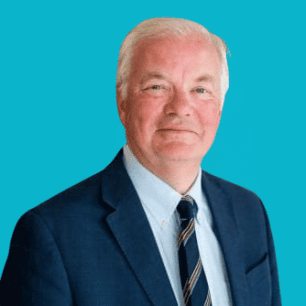 The image of a man from the shoulders up in a suit and tie, smiling into the camera with a bright background.