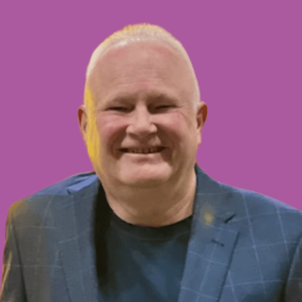 The image of a man from the shoulders up in a t-shirt and tie, smiling into the camera with a bright background.