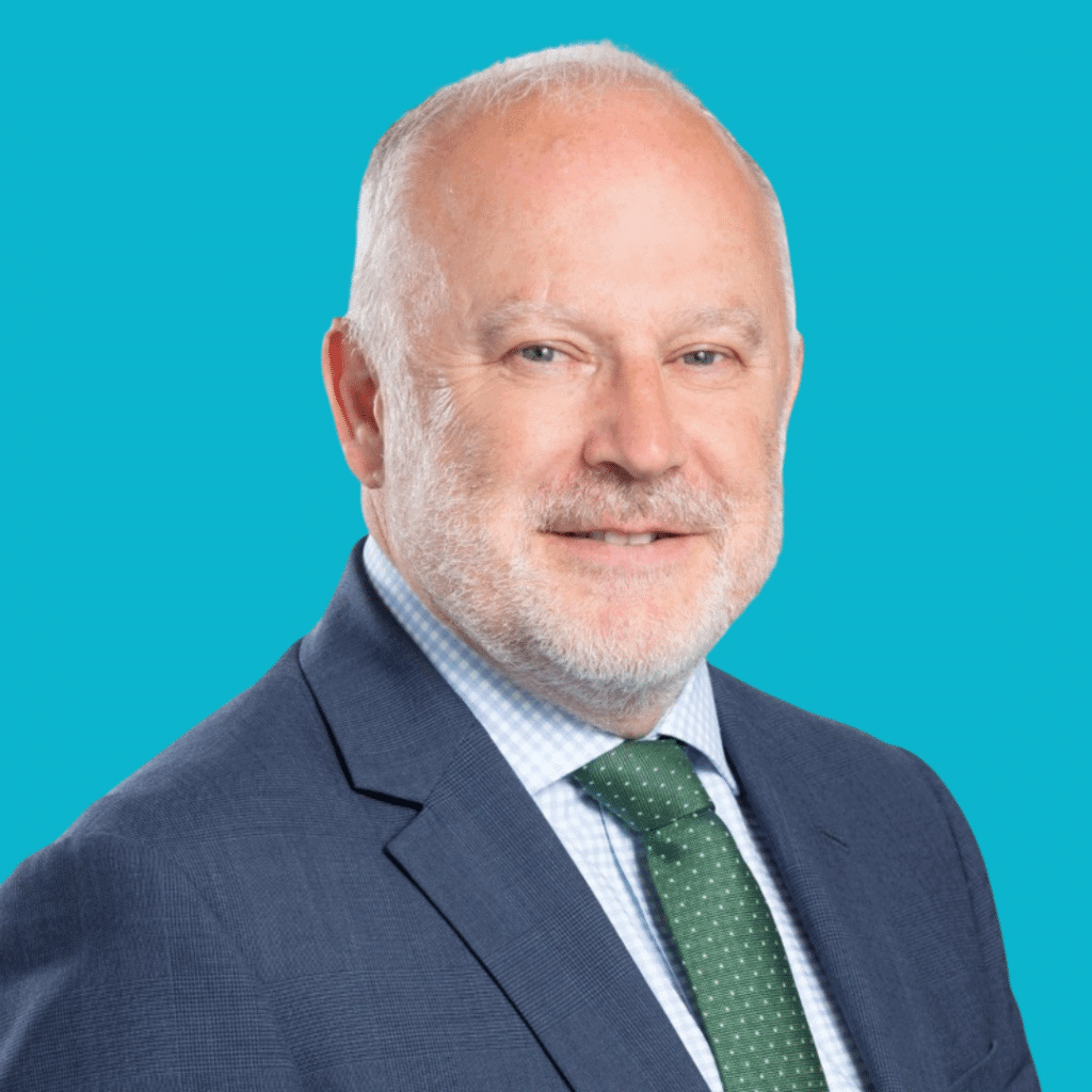 This picture is a of a man, from the shoulders up, dressed in a suit and tie. He's smiling into the camera on a bright background.