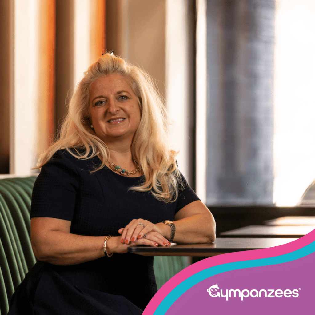 A smiling woman from the Gympanzees Commercial Property Committee leaning on a table in a modern indoor space, with Gympanzees branding in the corner.