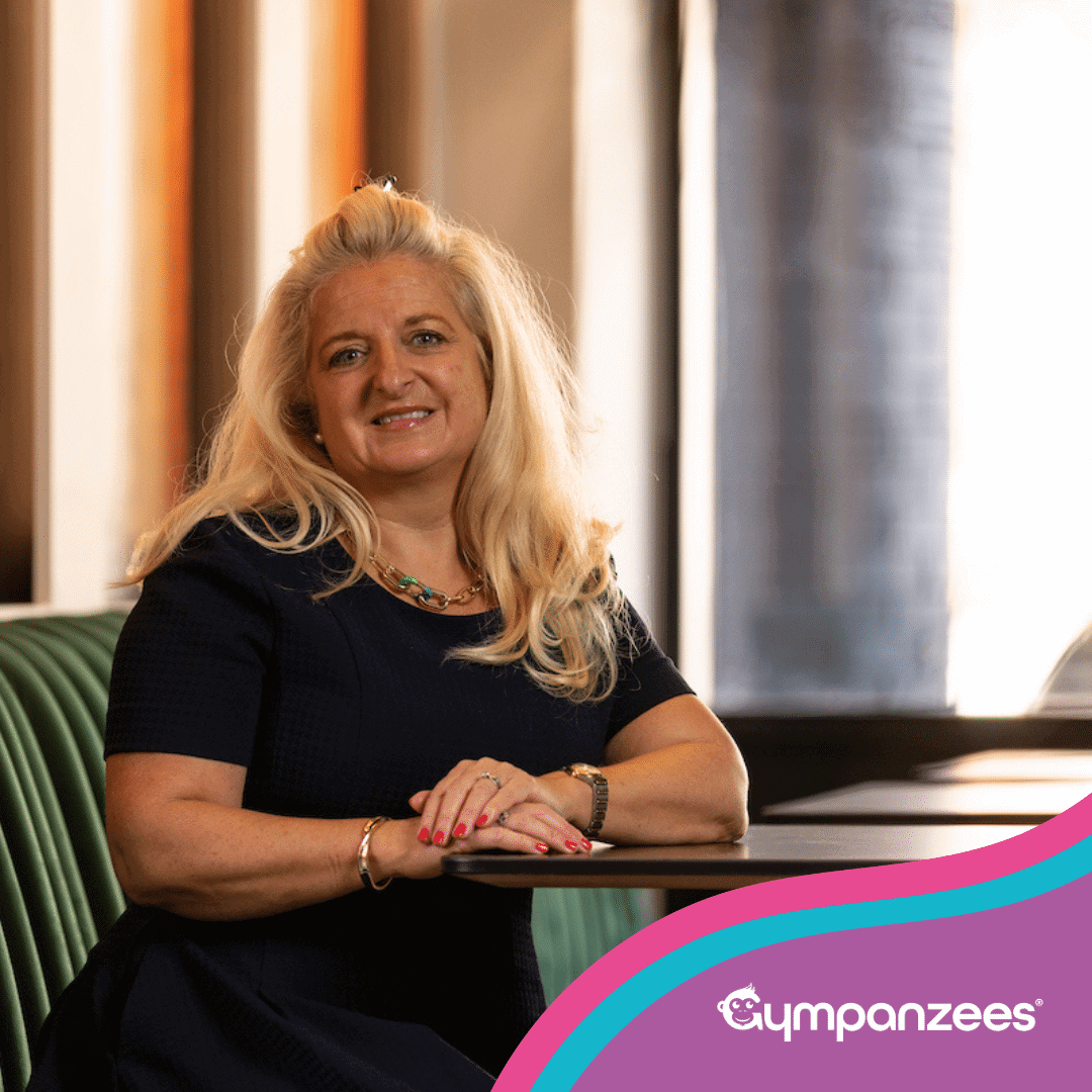 A smiling woman from the Gympanzees Commercial Property Committee leaning on a table in a modern indoor space, with Gympanzees branding in the corner.