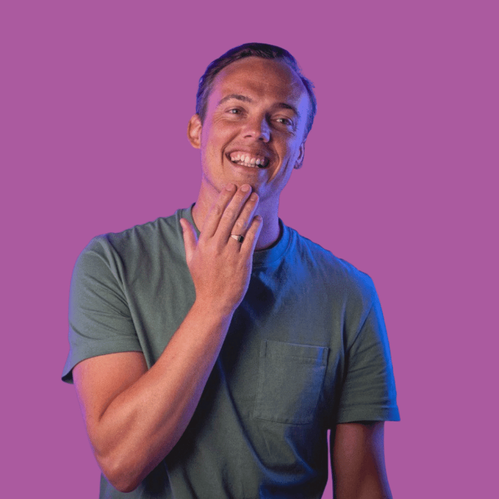 The image is of a man,holding his chin, in a dark t-shirt, smiling into the camera on a bright background.