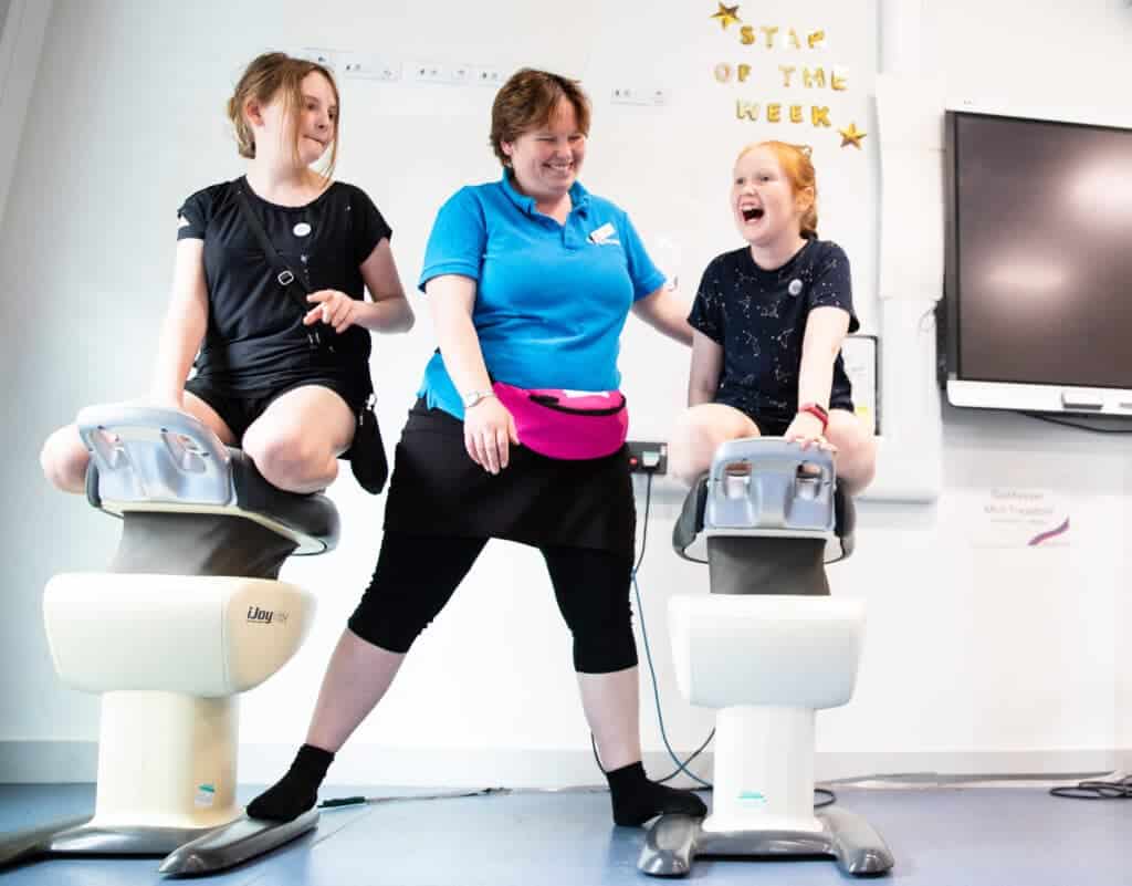 Image shows 3 people in the gym. An adult supervising 2 young children on horse riding simulator equipment. They are all smiling