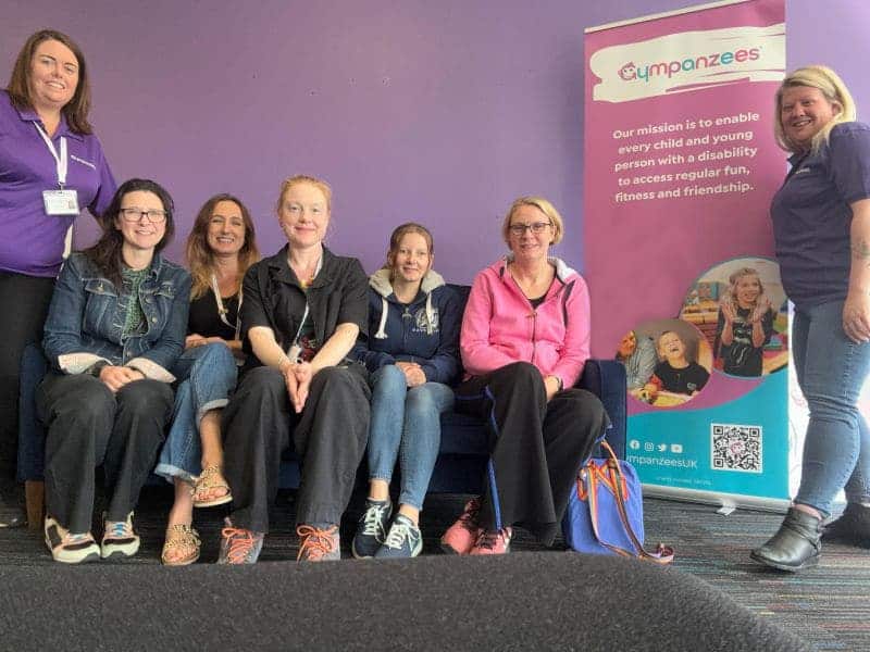 The image shows a group of individuals sat on a sofa smiling into the camera with two individuals, one stood at either end, in Gympanzees branded tops, with a branded Gympanzees banner to the right.