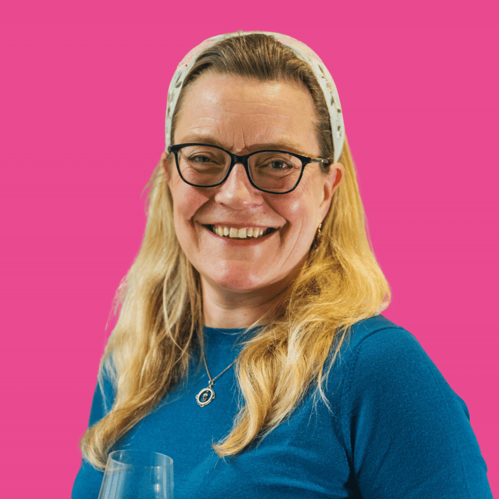 The image is of a lady with light long hair, a headband and glasses from the shoulders up smiling into the camera, on a bright background.