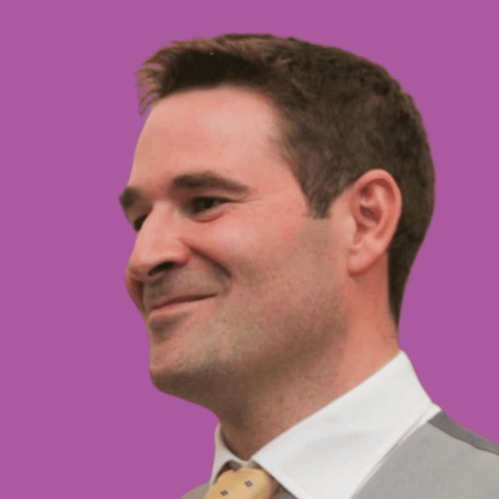 The image of a man from the shoulders up in a suit and tie, smiling away from the camera on a bright background.