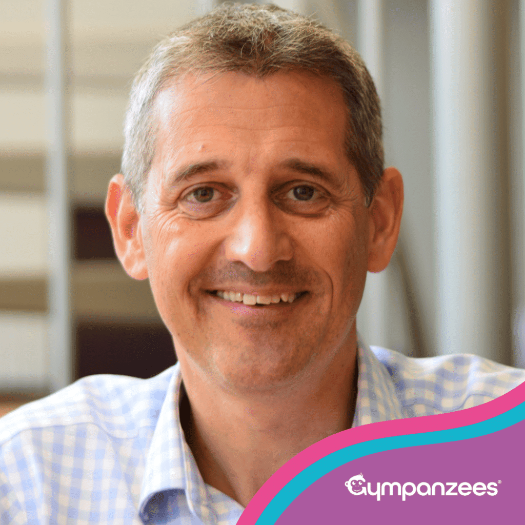 A smiling man with short gray hair and a light blue checkered shirt, standing in an office environment with blurred background elements. The image includes a colorful gradient overlay in the bottom right corner with the logo "Gympanzees" and a small monkey icon.