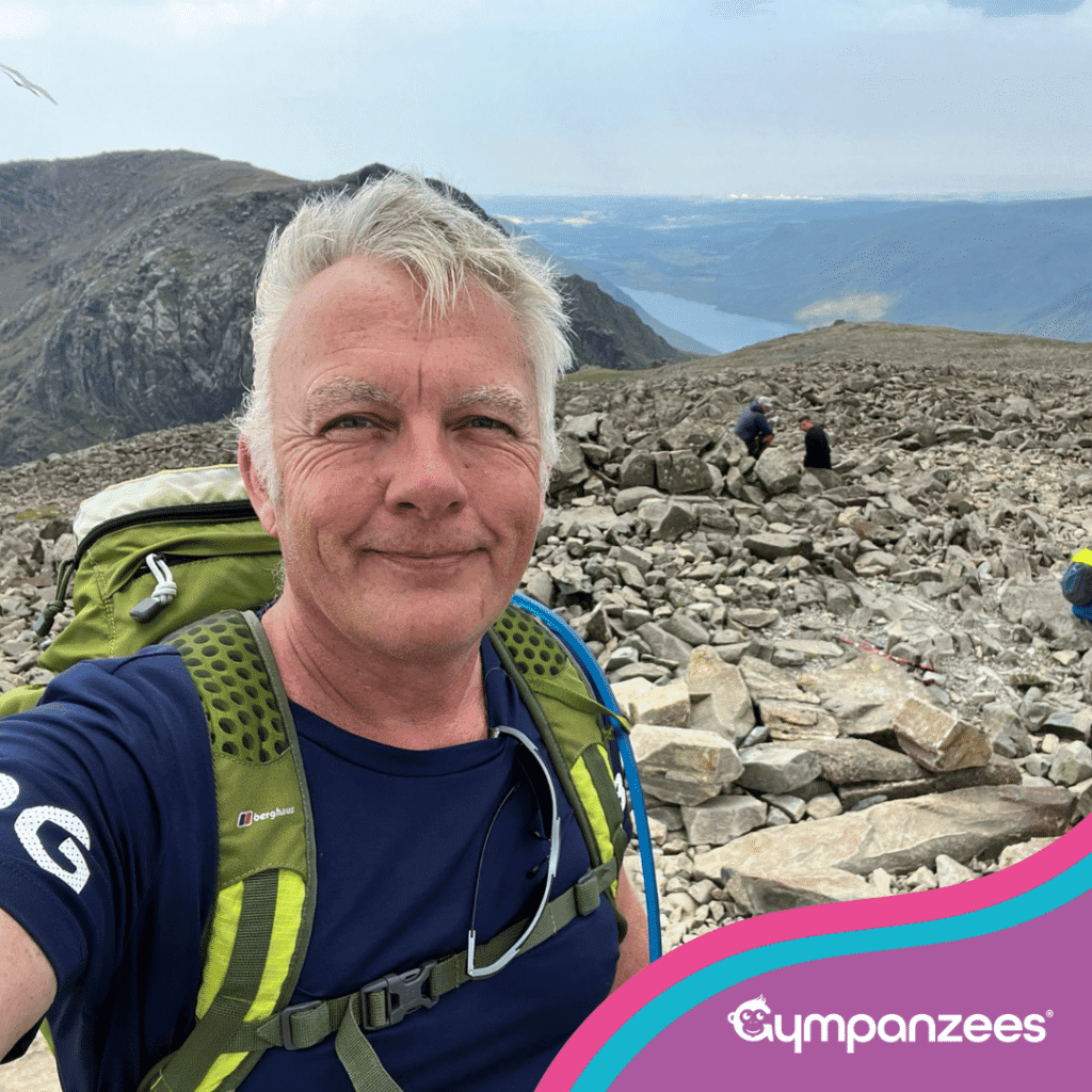 Person taking a selfie on a rocky mountain summit, wearing a backpack, with a wide landscape view of mountains and water in the background.