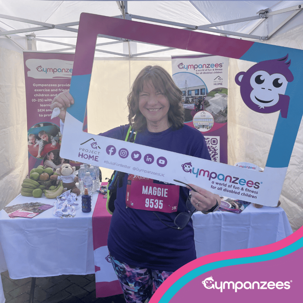 Person standing inside a Gympanzees event tent, smiling and holding a large branded photo frame, with information displays and refreshments visible behind them