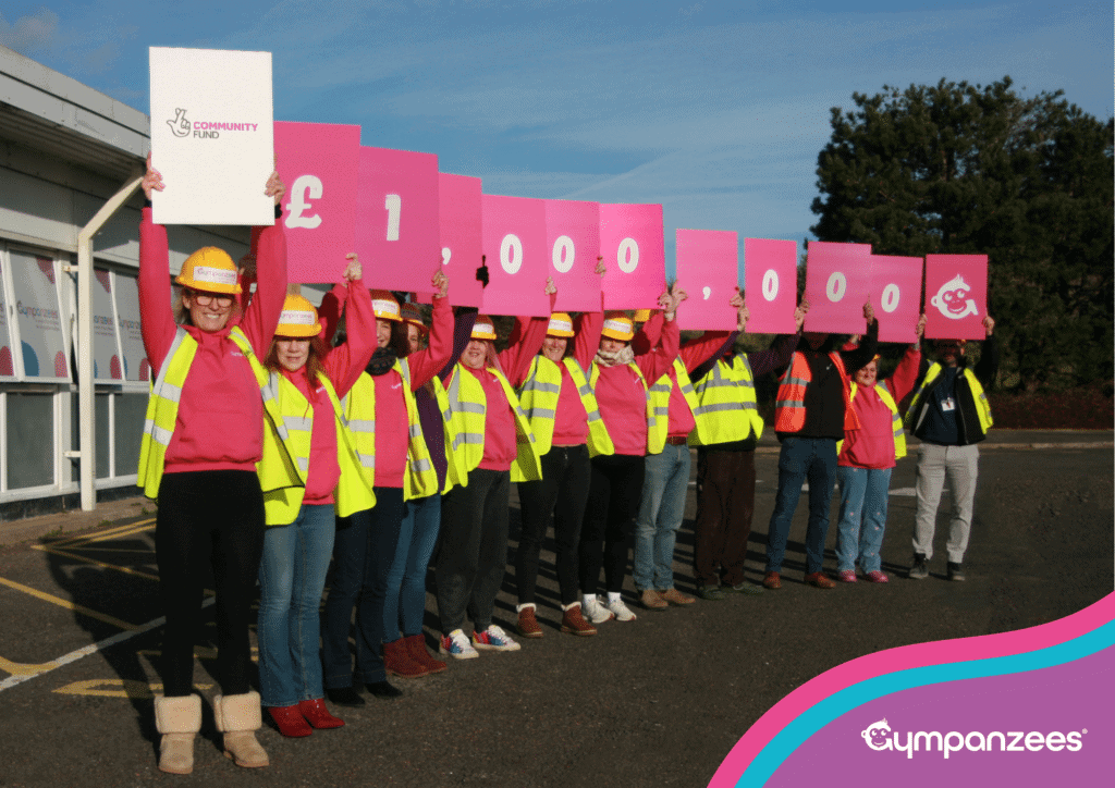 A group of people wearing high-visibility vests and pink tops stand outdoors holding large pink signs spelling out “£1,000,000,” with one sign showing The National Lottery Community Fund logo.