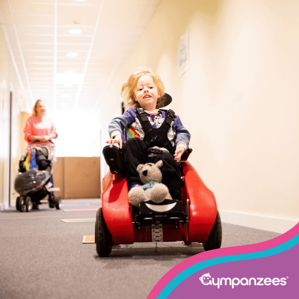 A young child using a powered mobility car in a hallway, with a soft toy on their lap, moving independently while an adult follows behind. Gympanzees branding is visible.