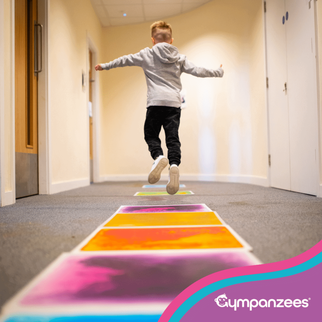 A child jumping along a colourful floor path in a hallway, mid-air with arms outstretched, taking part in an active play activity. Gympanzees branding is visible..