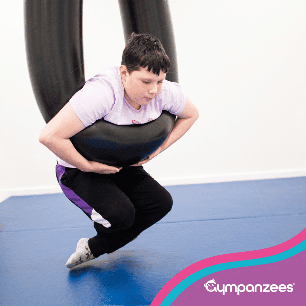 A child using a suspended inflatable swing, holding on while lifting their body off the floor in a supported movement activity. Gympanzees branding is visible.
