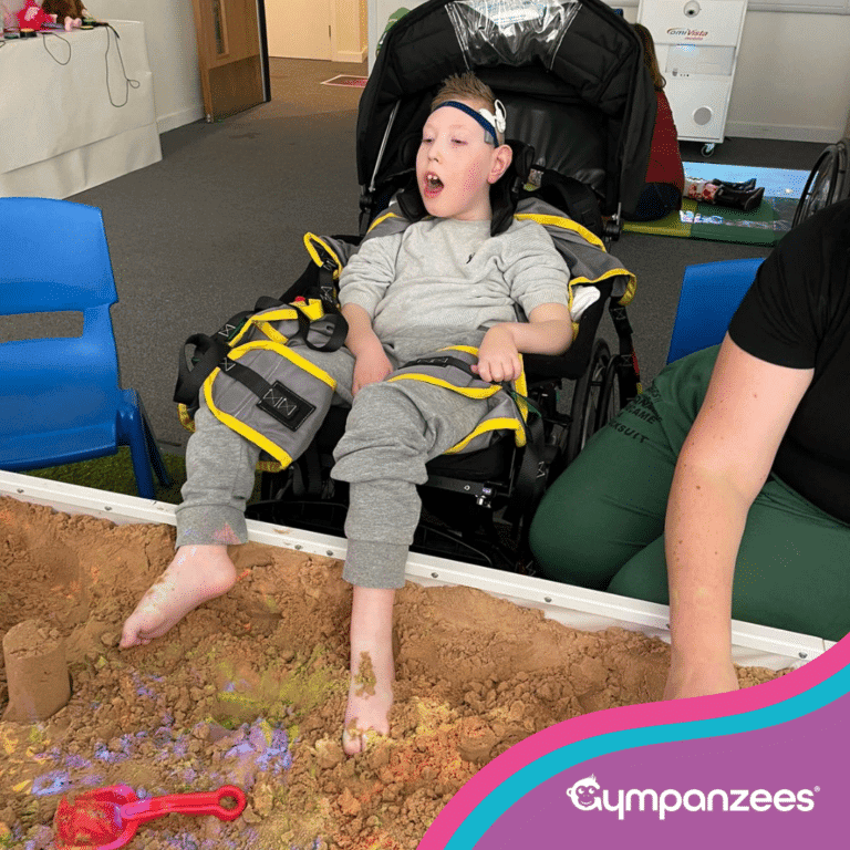 A child sits supported in a specialist wheelchair, exploring a sand play tray with their feet. Their toes are buried in the sand while colourful light reflections scatter across the surface. Gympanzees branding appears in the corner.