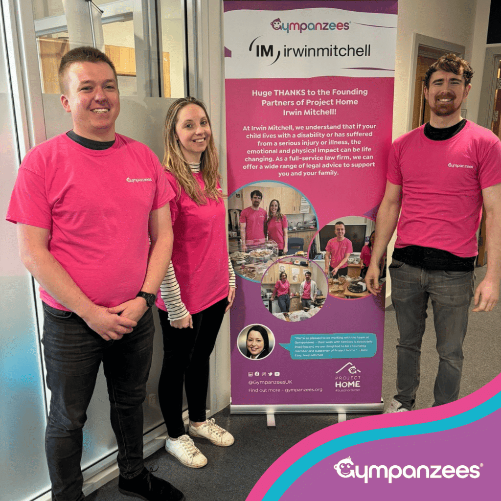 Three people stand smiling in front of a banner with the logos of Gympanzees and Irwin Mitchell. They are wearing matching pink t-shirts.