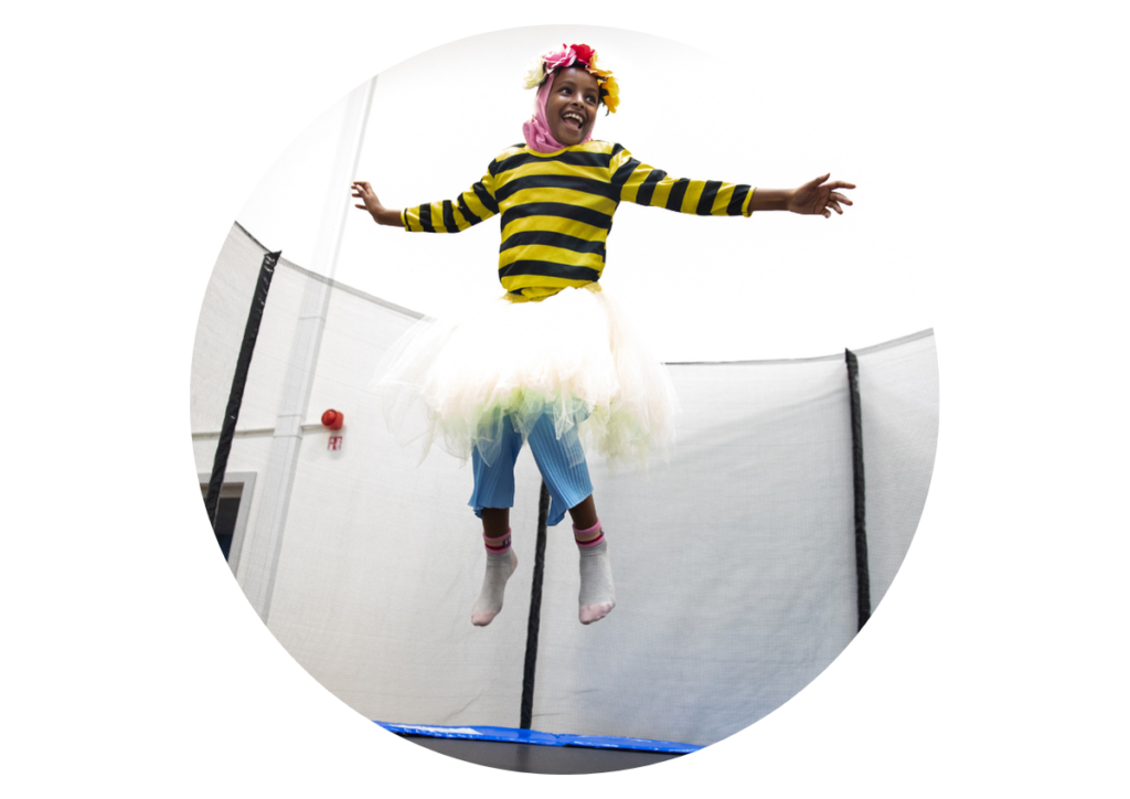 The image is of a girl high up in the air, bouncing on a trampoline. She is wearing a colour top, flower crown, flowing skirt and leggings.