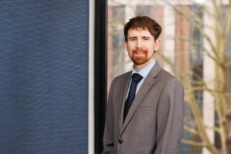 The image is of a man with a suit on, short dark hair, facial hair facing the camera smiling.