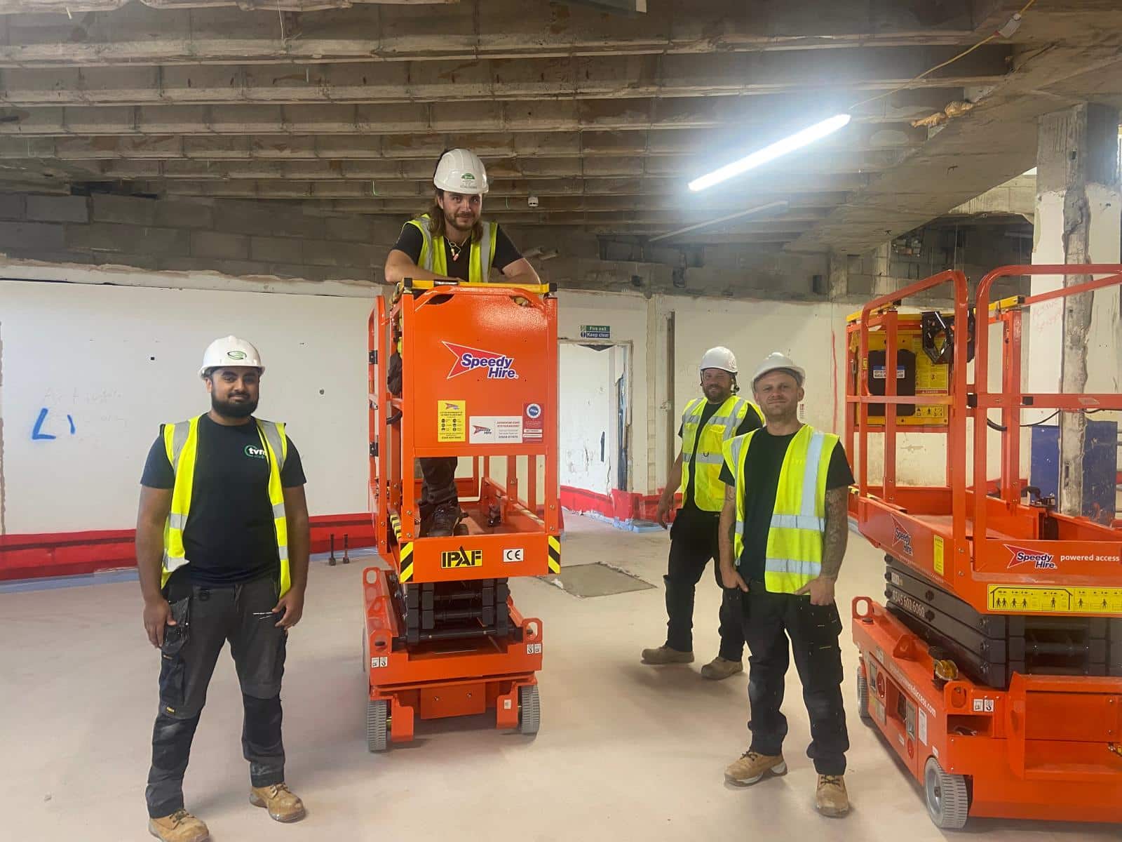 Four construction workers wearing hard hats and high visibility vests stand inside the Gympanzees Centre during the build. Two orange access platforms are positioned in the space, with exposed ceiling beams and unfinished walls visible in the background.