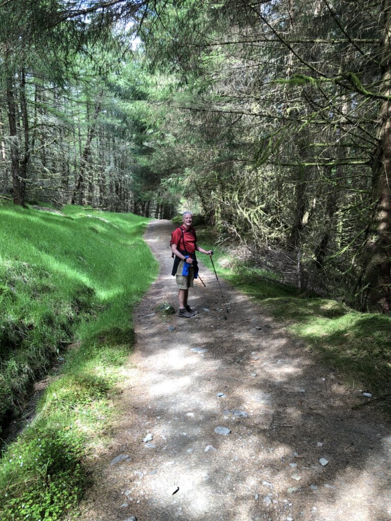 A man wearing walking gear and holding two walking poles stands on a countryside trail, surrounded by grassy hills and woodland