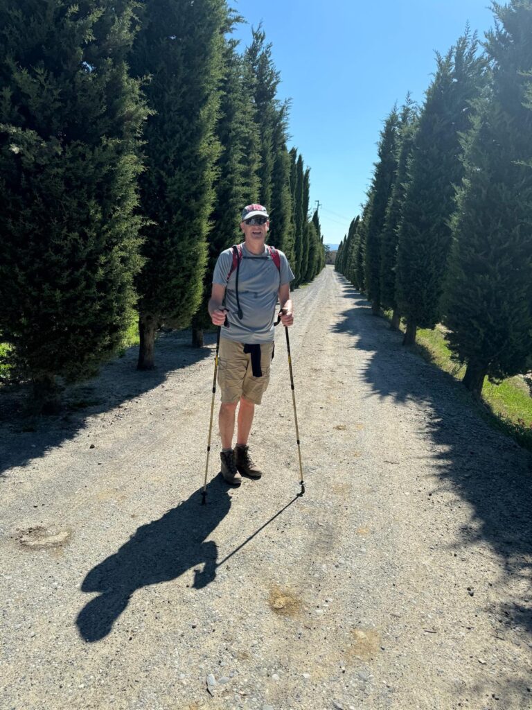 A man wearing walking gear and holding two walking poles stands on a countryside trail, surrounded by grassy hills and woodland