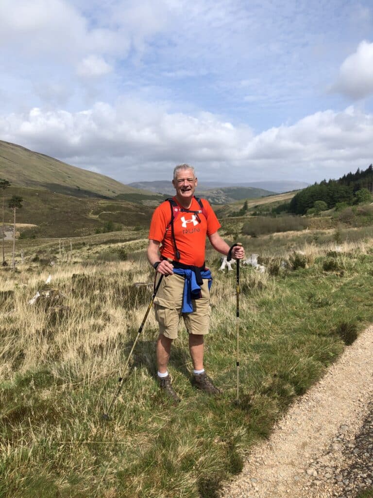 A man wearing walking gear and holding two walking poles stands on a countryside trail, surrounded by grassy hills and woodland