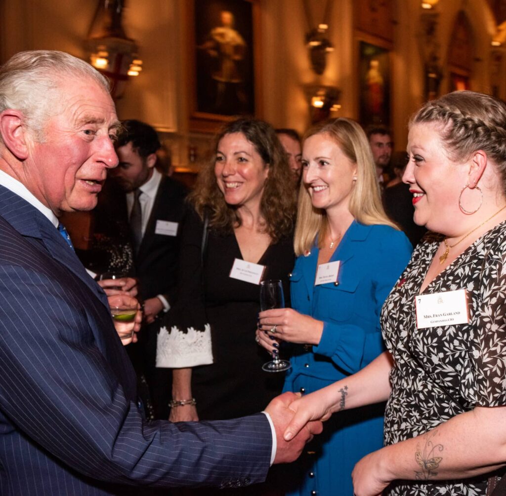 Fran Garland is pictured at an event, smiling as she shakes hands with the King. Several other guests stand nearby, smiling and chatting in a warmly lit room.