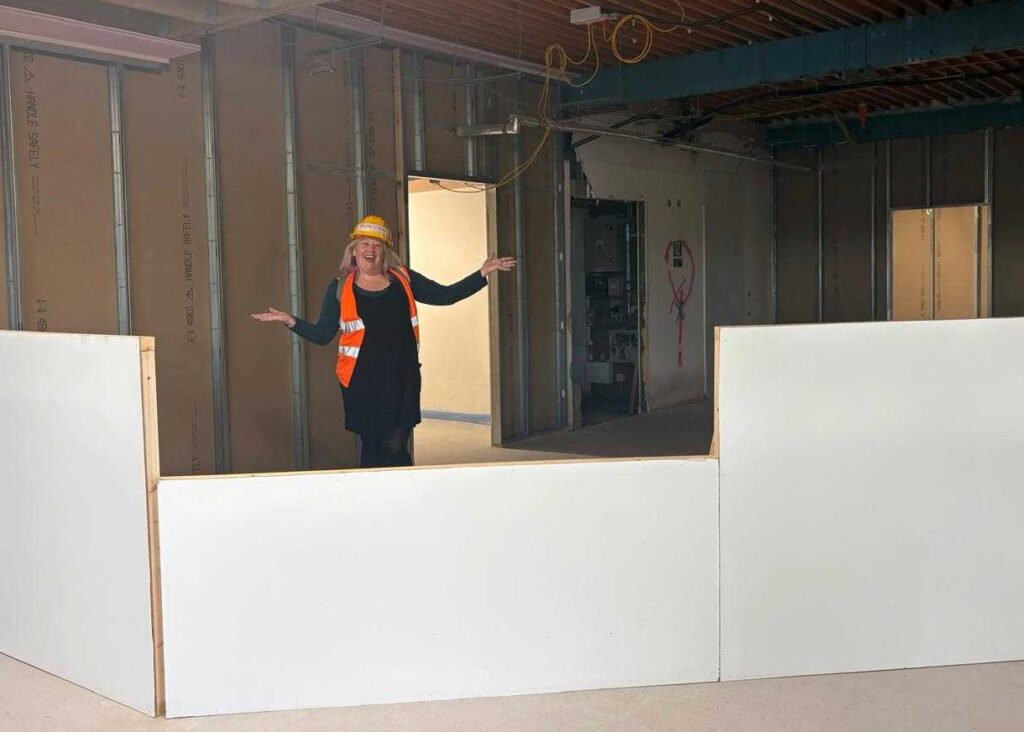 The image is of a large building space with a mock up desk at the front with a lady with her hands in the air smiling, with a hard hat and high vis on.