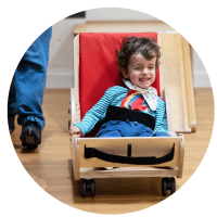 The image is of a young boy sat, with a look of excitement, in a wooden wheelable cart coming toward the camera.