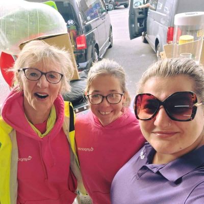 The image of three women with brightly coloured tops on, all wearing glasses apart from the on the right in sunglasses, with a couple of vans in the background.