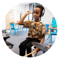 Image of a young boy with a dark tree patterned t-shirt sat on a stool in front of some drums with headphones on and a drumstick in one hand and his other hand holding the headphones to his head. This is in a classroom setting in the background.